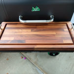 Wooden cutting board on a grill with a visible brand logo.