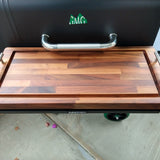 Wooden cutting board on a grill with a visible brand logo.