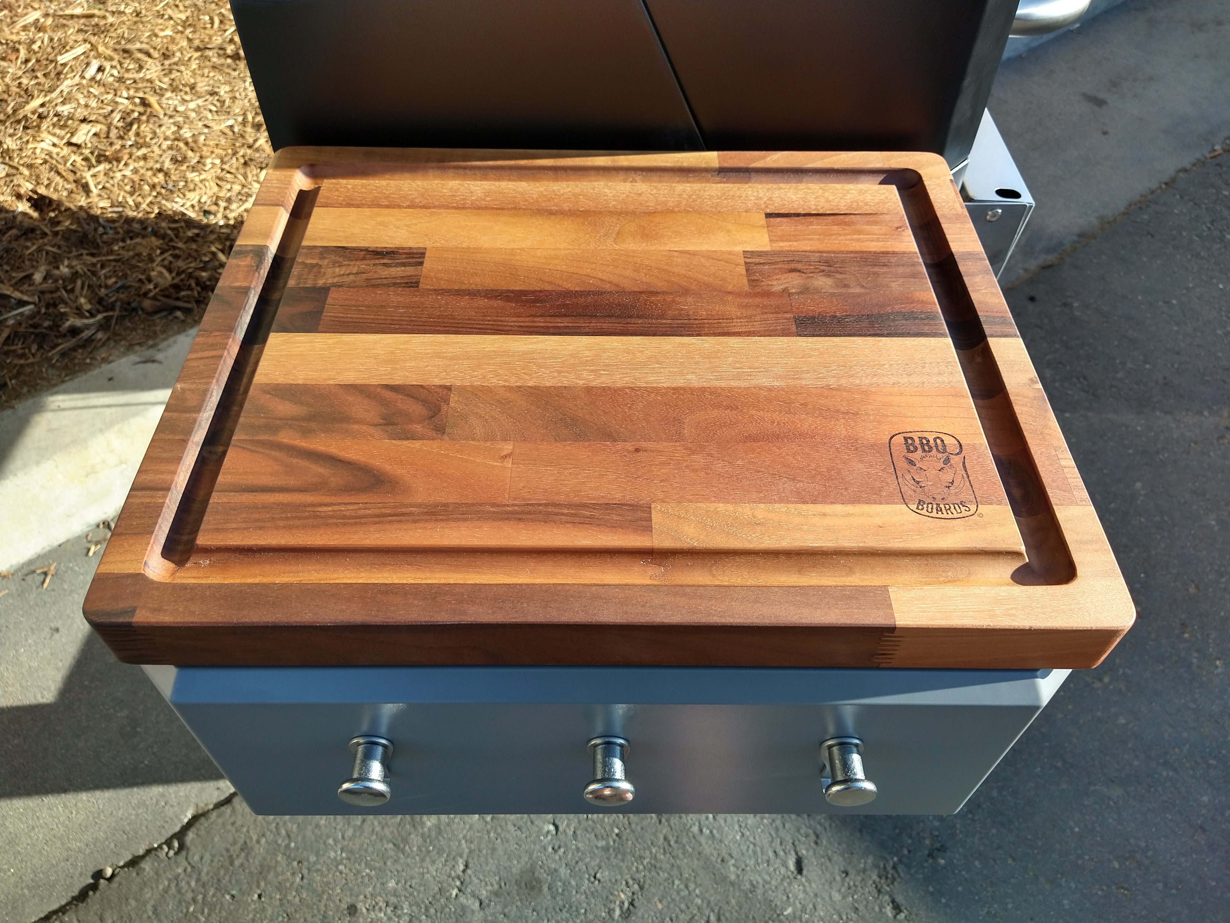 Wooden cutting board on a grill with a brand logo.