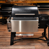 A Traeger pellet grill with a black lid and stainless steel lower drawer and legs, shown on a wood floor with other grills in the background.