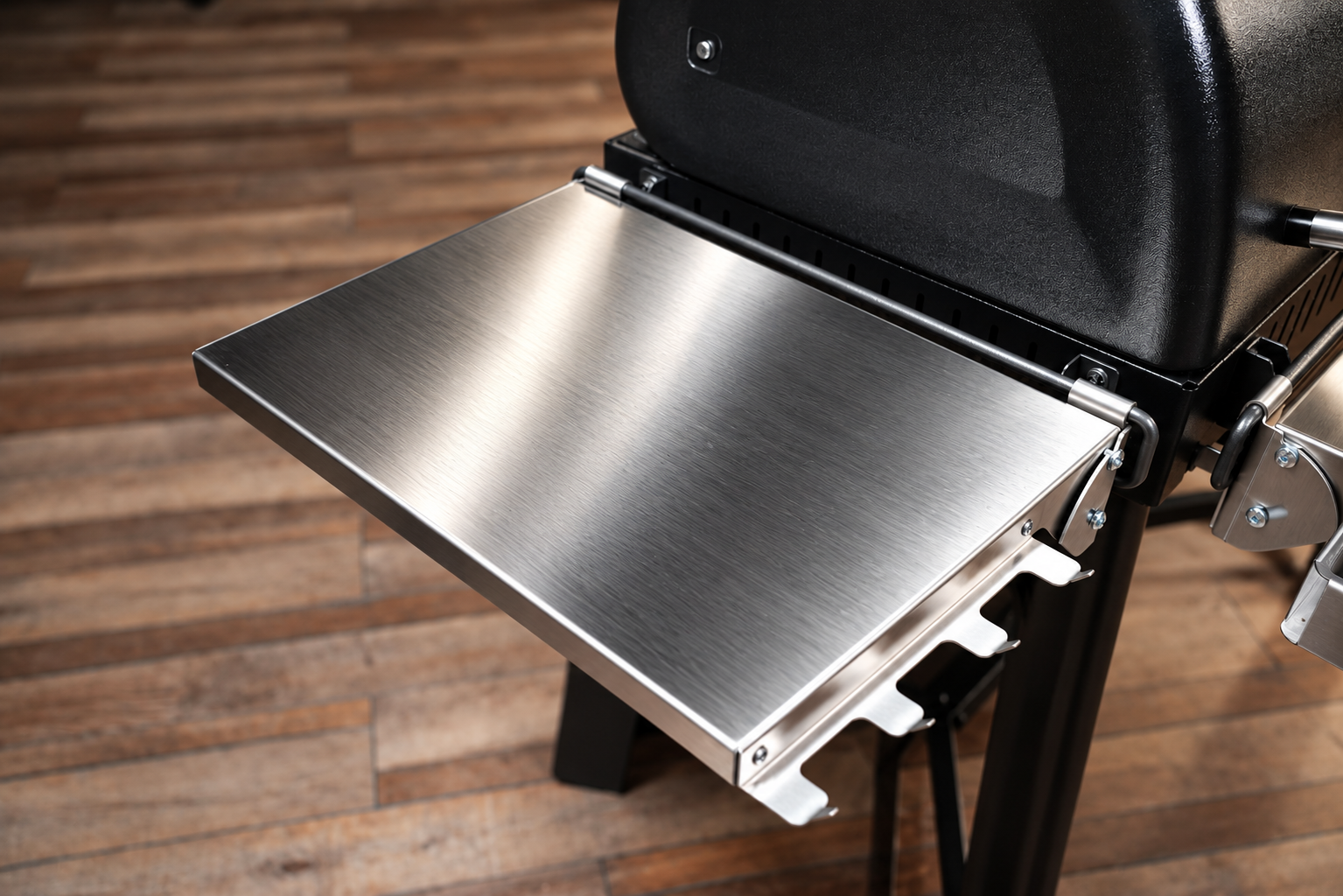 Close-up of a brushed stainless steel side table fold-out shelf attached to a black grill, set against a wooden plank floor.