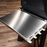 Close-up of a brushed stainless steel side table fold-out shelf attached to a black grill, set against a wooden plank floor.