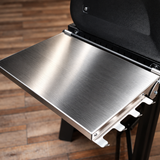 Close-up of a brushed stainless steel side table fold-out shelf attached to a black grill, set against a wooden plank floor.